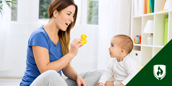 Photo of a female caregiver holding a rubber duck in front of an infant sitting upright and locking eyes on the toy