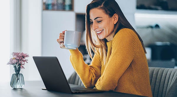 smiling woman sitting at a desk looking at a laptop and holding a cup