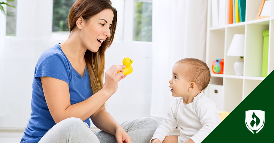 Photo of a female caregiver holding a rubber duck in front of an infant sitting upright and locking eyes on the toy