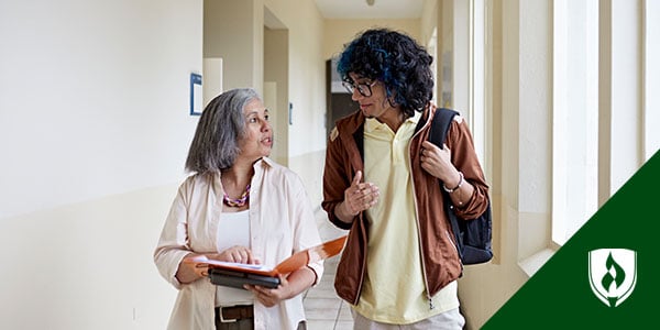 An older woman talks to a young man with blue in his hair as they walk down a hall.