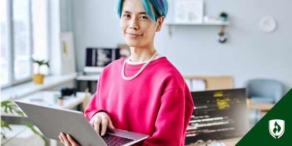 A business major with blue hair and a computer stands in their home office