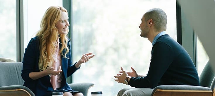 Photo of professionally-dressed woman and man having a sit down conversation in an office.